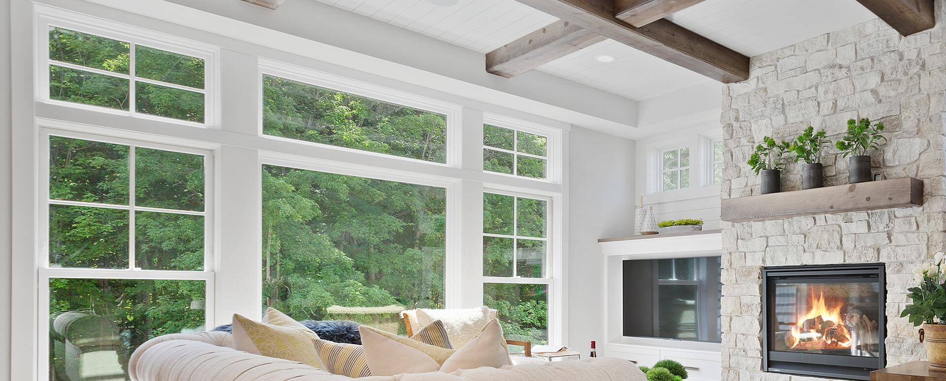 Transom windows above large windows in a large living area with stone fireplace and a clear view of the outside, with trees beyond.