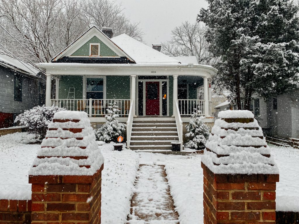 Cottage-style home in the heavy snow. Green siding and large front porch.