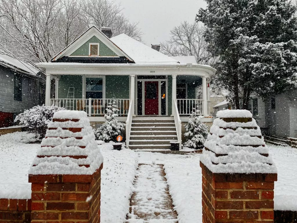 Cottage-style home in the heavy snow. Green siding and large front porch.