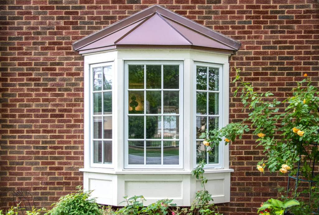 Bay window with copper roof on traditional brick home with flowers below and yellow roses extending out onto the window in springtime